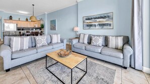 A modern living room with two grey sofas, a coffee table, and a kitchen in the background. - Ocean Villa 2004 (Panama City Beach)
