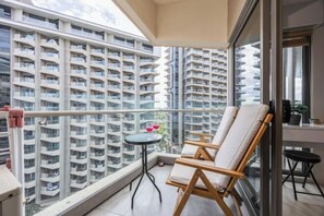 Uma varanda com vista para edifícios altos, uma mesa redonda pequena com dois copos e duas cadeiras de madeira.