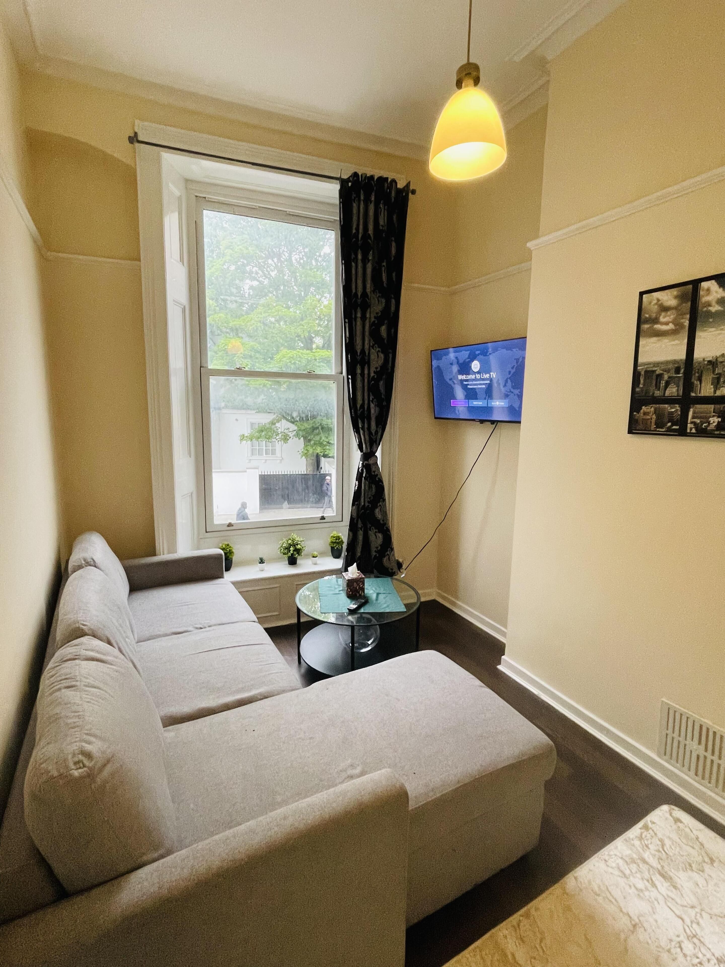 Ein gemütliches Wohnzimmer mit einer Couch, einem kleinen runden Tisch, einem an der Wand montierten Fernseher und einem Fenster mit Vorhängen.