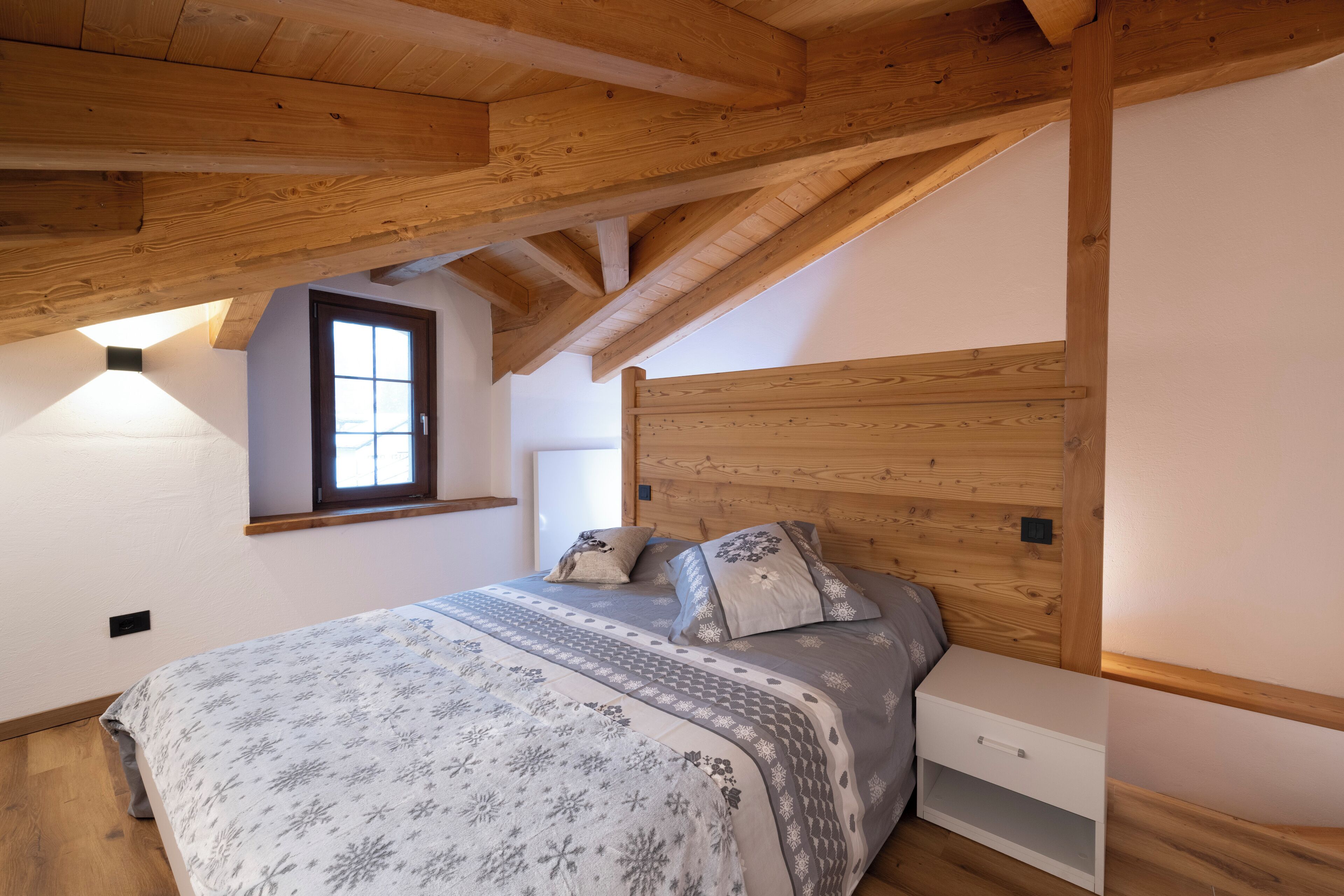 A bedroom with a wooden ceiling, a bed with patterned bedding, a nightstand, and a window.