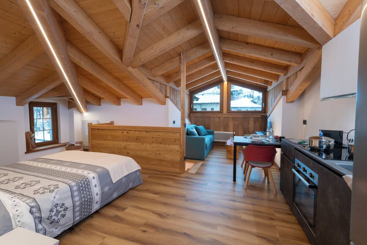 A bedroom with a wooden ceiling, a bed, a sofa, a dining table, and a kitchen area.