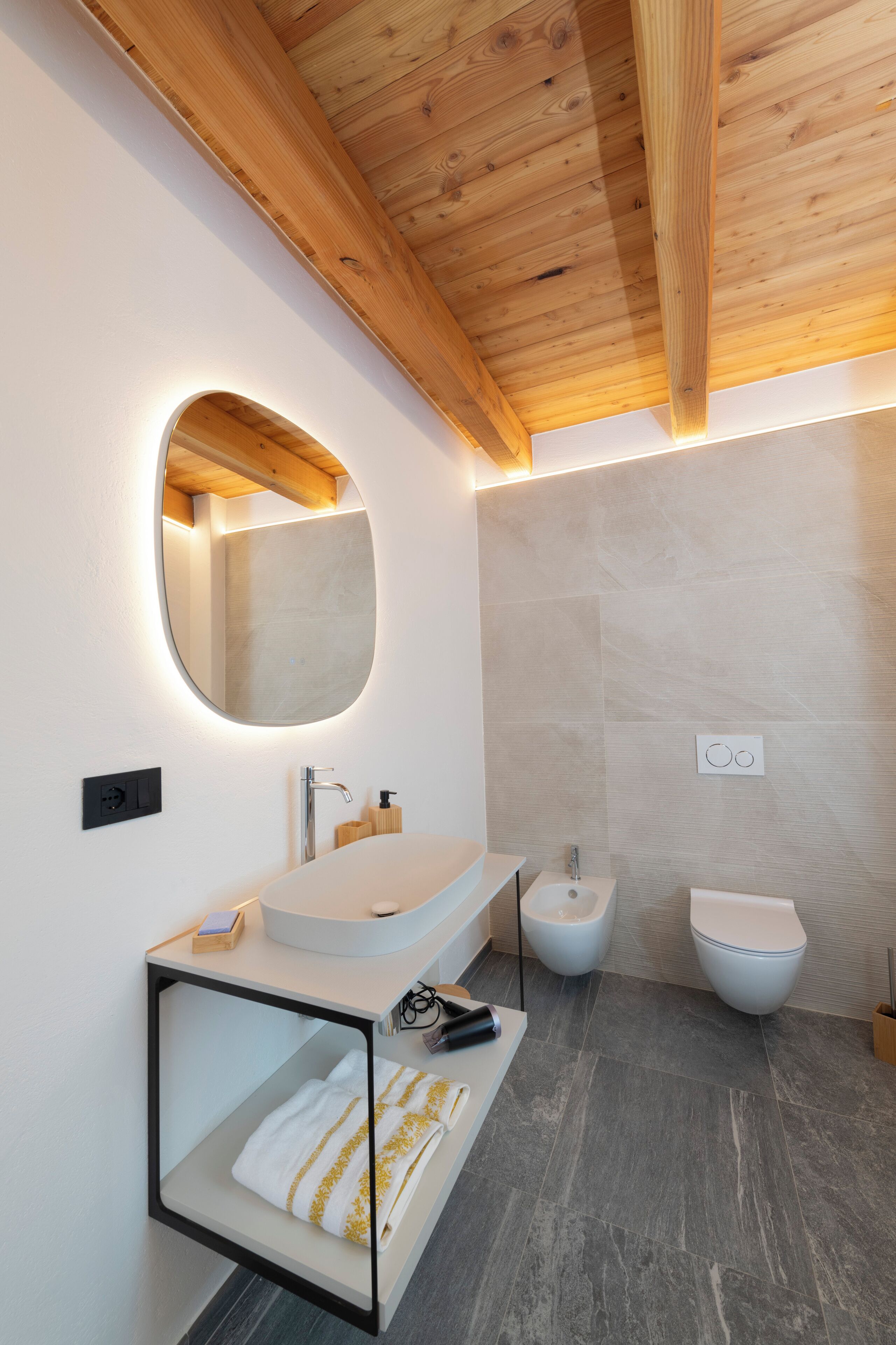 A modern bathroom with a wooden ceiling, a round mirror, a white sink, and two toilets.