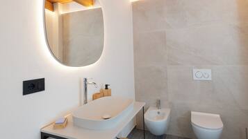 A modern bathroom with a wooden ceiling, a round mirror, a white sink, and two toilets.