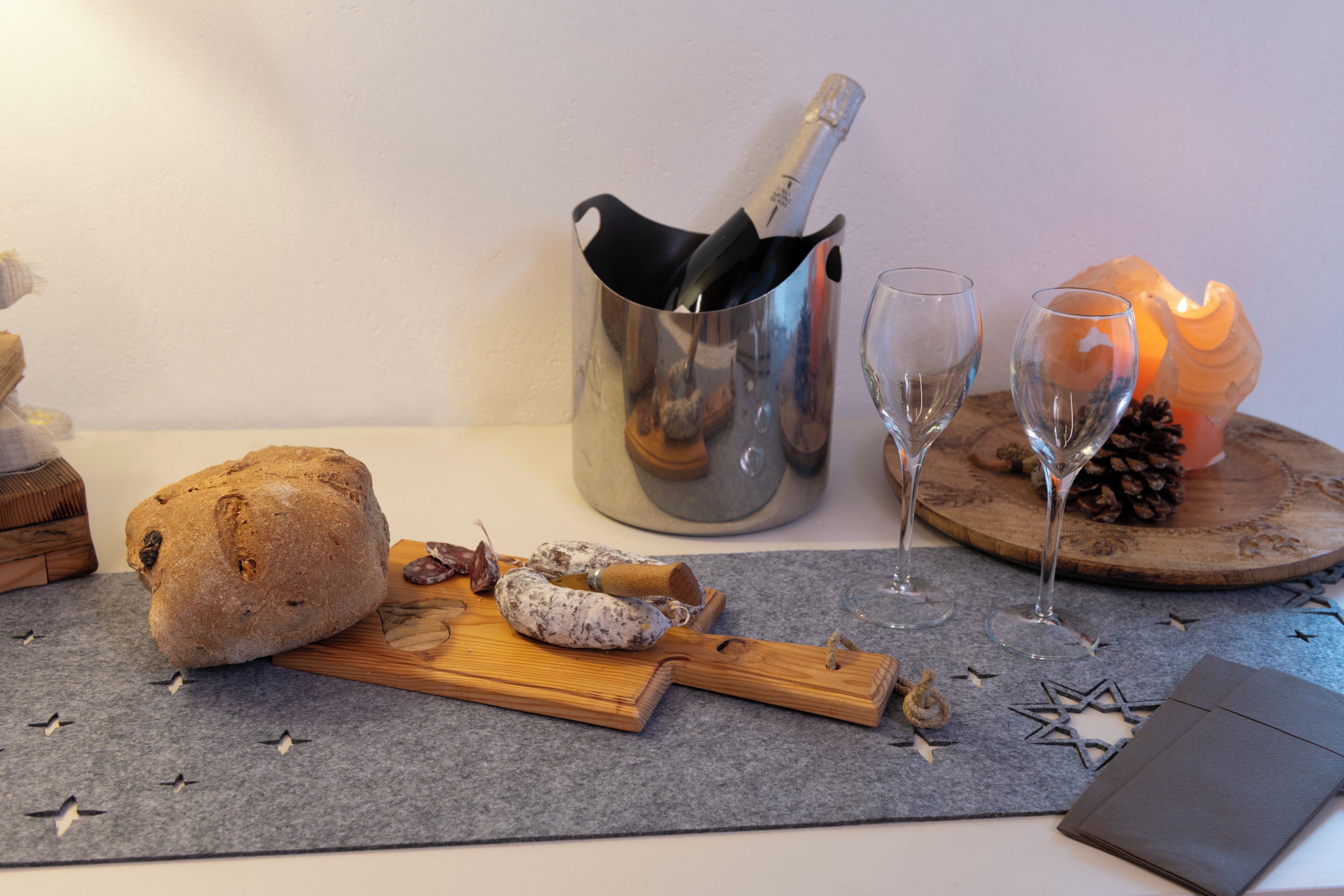Ein Tisch mit Brot, einem Holzschneidebrett, einer Champagnerflasche, zwei Weingläsern und einer Kerze auf einer Filzunterlage.