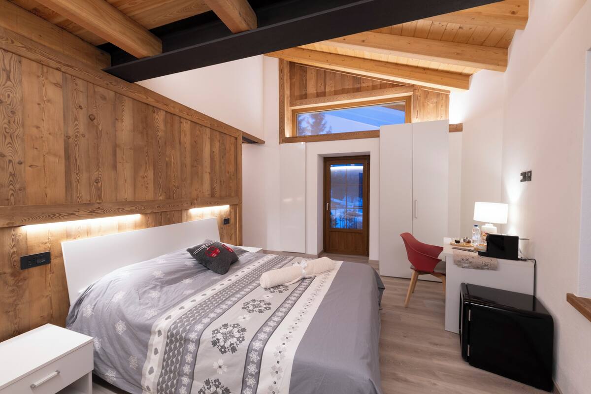 A modern bedroom with a wooden ceiling, a bed with patterned bedding, a desk with a lamp, and a red chair.