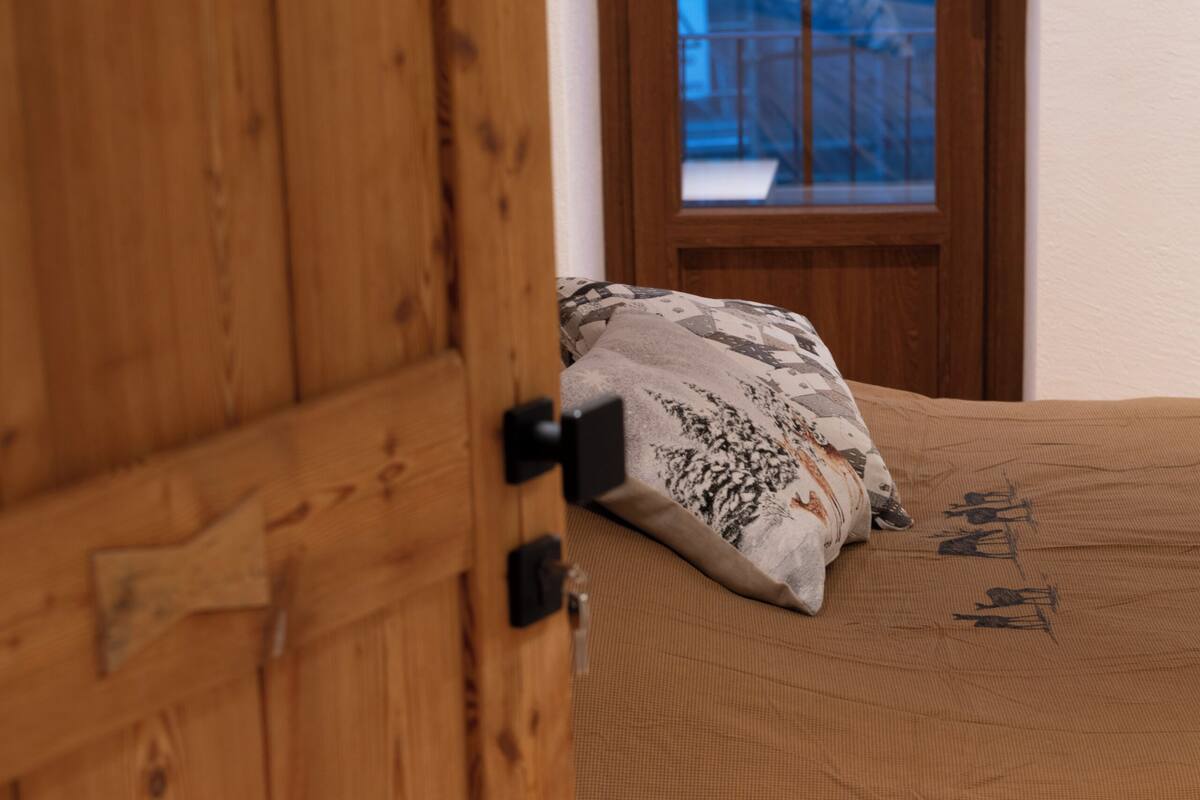 A wooden door with a black handle, a bed with a brown bedspread and patterned pillows, and a window with a view of a balcony.