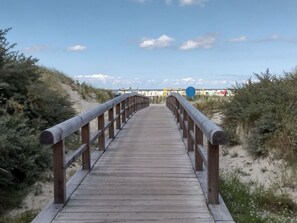Une promenade en bois qui mène à une plage bordée de dunes de sable et dotée d’une végétation clairsemée.
