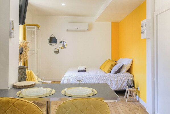Quarto de hotel com uma cama, uma mesa de jantar posta com pratos e talheres, um banquinho pequeno e um espelho na parede.