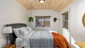 A bedroom with a bed, a nightstand, a lamp, a window with blinds, and a hanging plant.