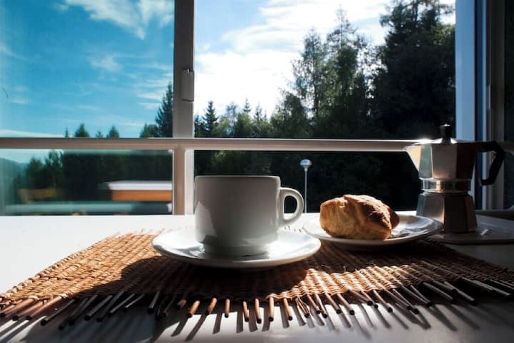 Une tasse de café et une pâtisserie sur une table près d’une fenêtre donnant sur des arbres.