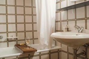 Une salle de bain avec un lavabo blanc, un miroir, un rideau de douche et un plateau en bois sur lequel se trouve un verre de vin.