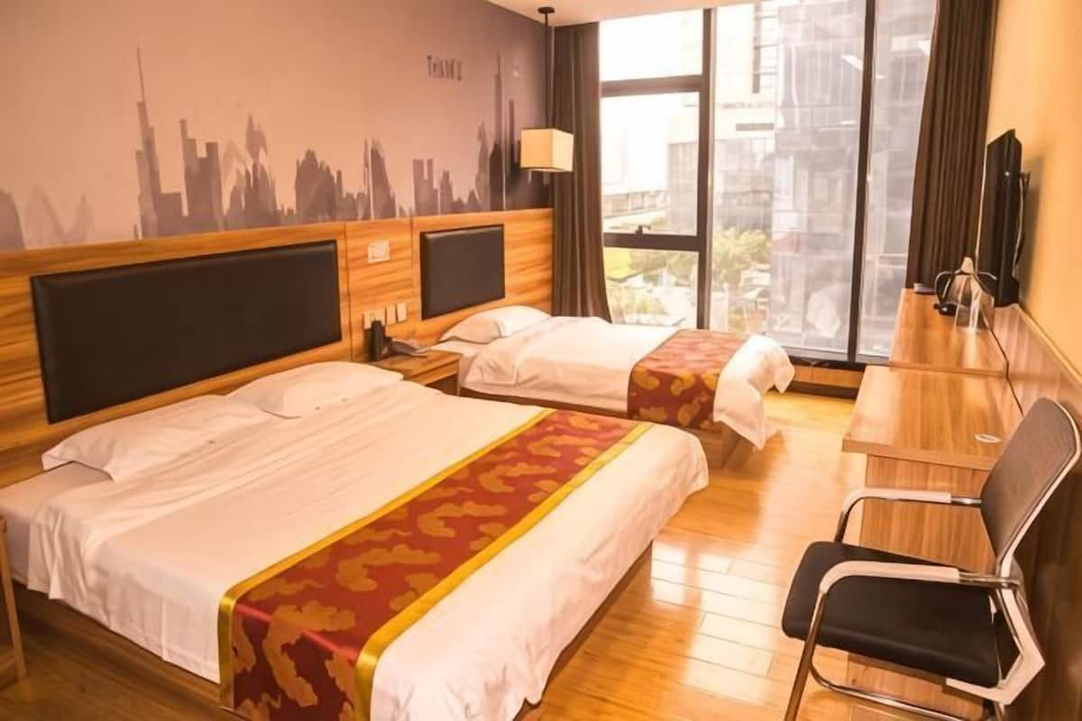 Ein Hotelzimmer mit zwei Betten, einem Schreibtisch, einem Stuhl, einem Fernseher und einem Wandbild mit Stadtansicht.