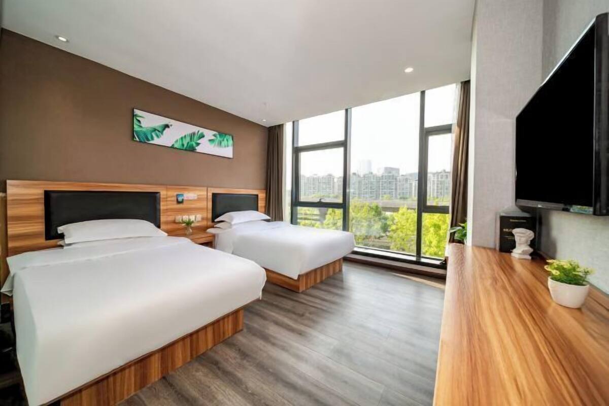 Ein Hotelzimmer mit zwei Betten, einem großen Fenster mit Blick auf die Stadt, einem Flachbildfernseher und einem Holztisch mit Pflanze.