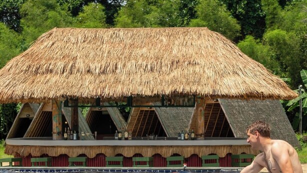 A swimming pool with two people on inflatable floats, a thatched-roof structure in the background, and lush greenery. - Slumber Resort Koh Chang (Ko Chang)