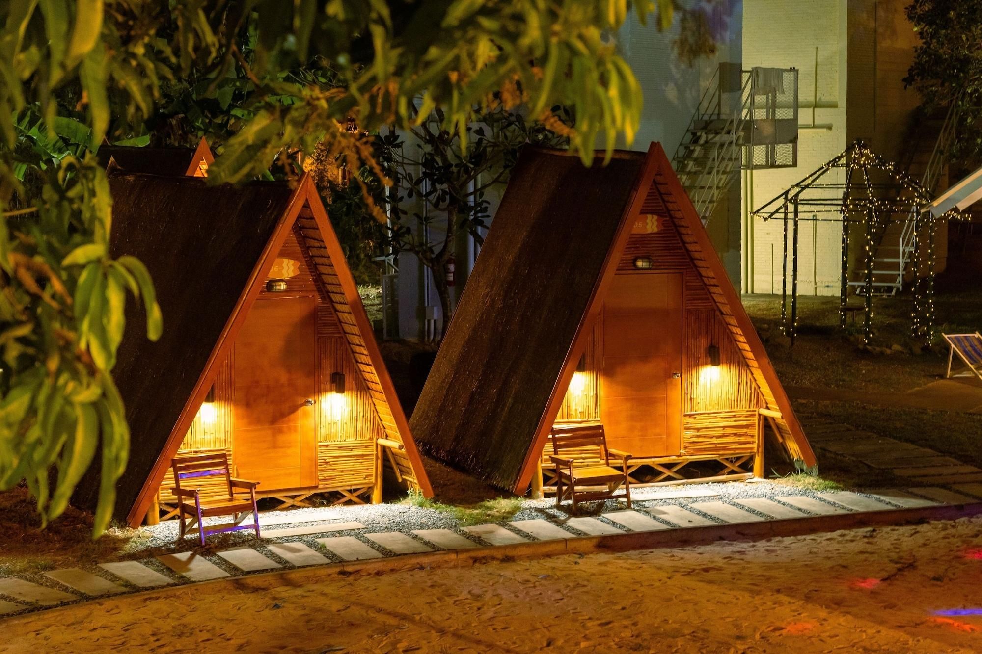 Two wooden A-frame cabins with interior lighting, a stone pathway, and a staircase leading to a building in the background.