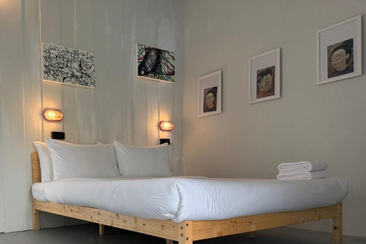 A bed with white bedding, a wooden headboard, and a wall-mounted light fixture.