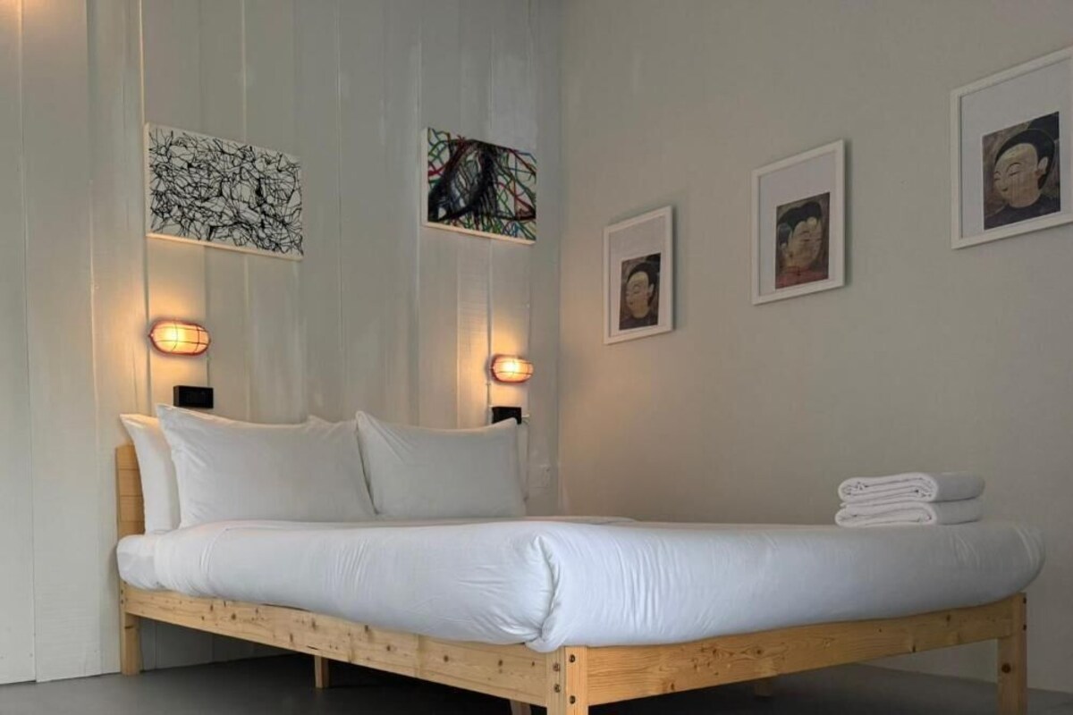 A bed with white bedding, a wooden headboard, and a wall-mounted light fixture.