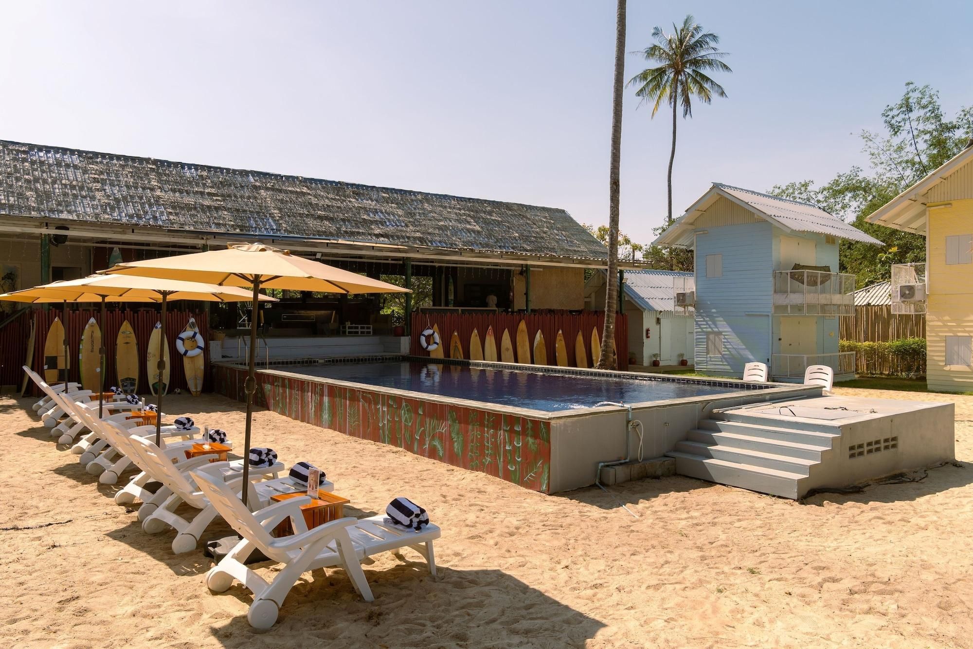 Uma área à beira da piscina com espreguiçadeiras e guarda-sóis, piscina e um edifício com telhado inclinado.