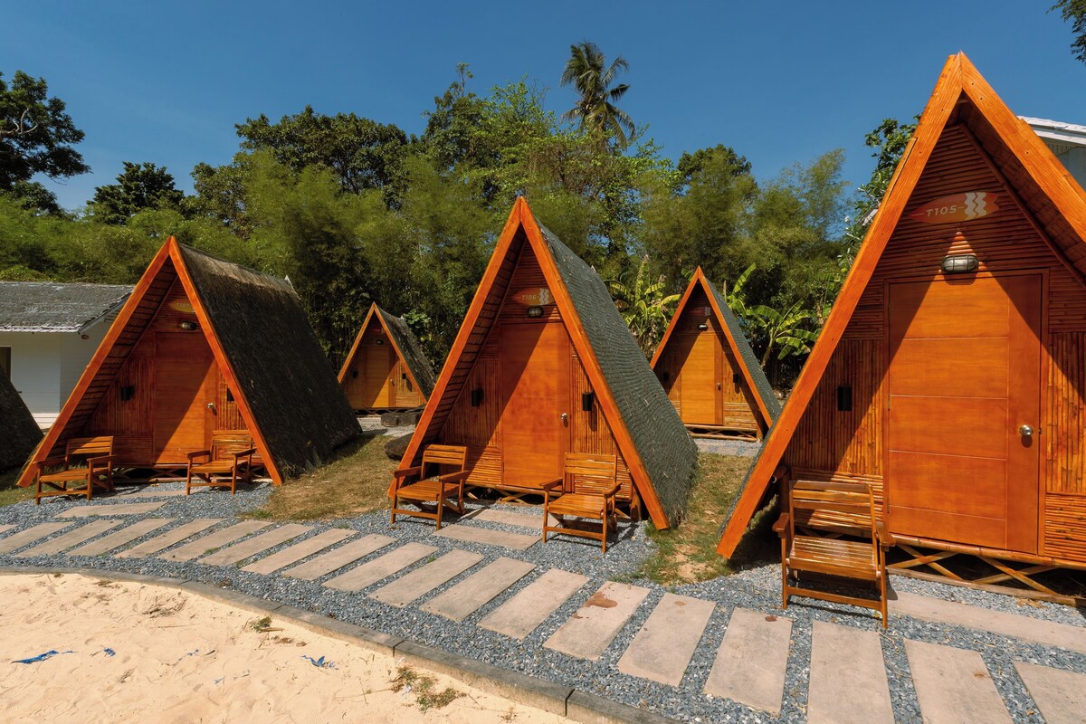 A row of wooden triangular cabins with built-in seating and a thatched roof, situated on a gravel path with a beach in the background.