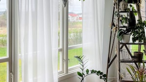 Huoneessa on suuret ikkunat, puinen kirjahylly, jossa on ruukkukasveja, ja ikkunasta avautuu näkymä punakattoiseen taloon.