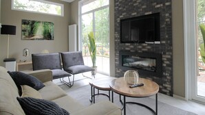A modern living room with a sofa, chairs, a coffee table, a fireplace, and a TV mounted on a brick wall. - Modern Holiday Home With Decorative Fireplace (Hoenderloo)