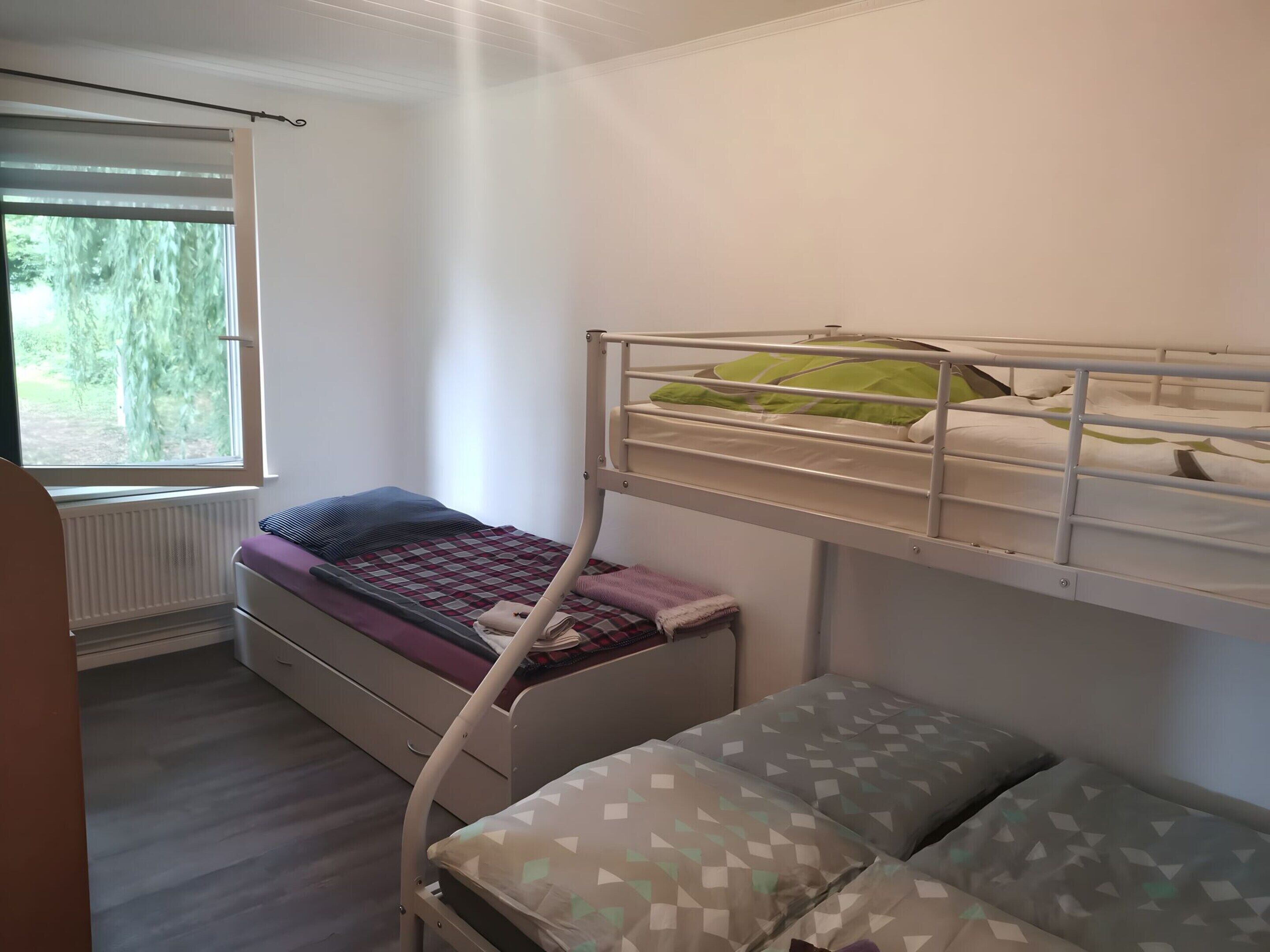 A bedroom with a bunk bed, a single bed, a window with blinds, and a radiator.