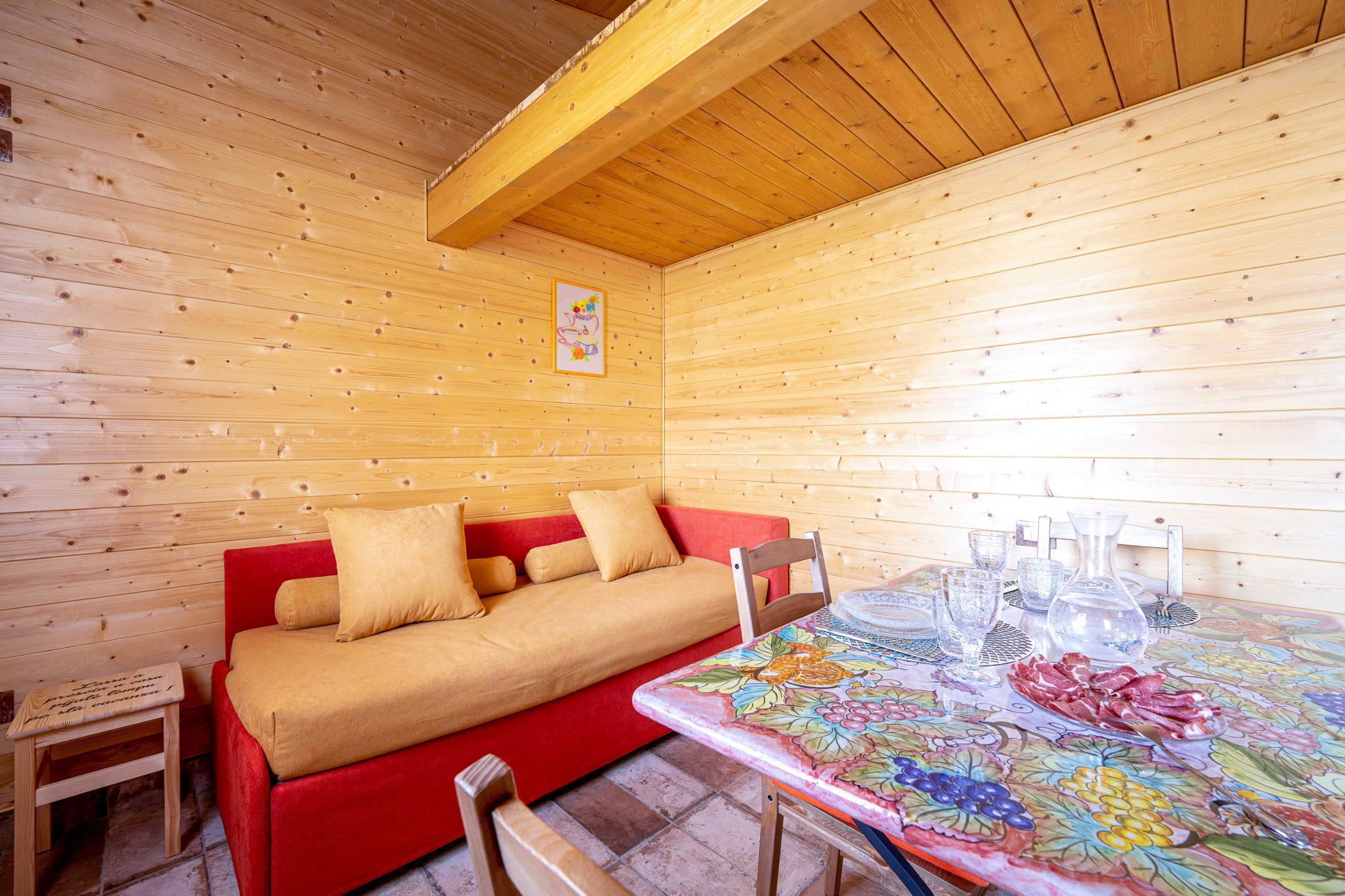 Ein Holzhütten-Innenraum mit einem roten Sofa, einem kleinen Tisch und einem Esstisch mit Gläsern und einer Obstplatte.