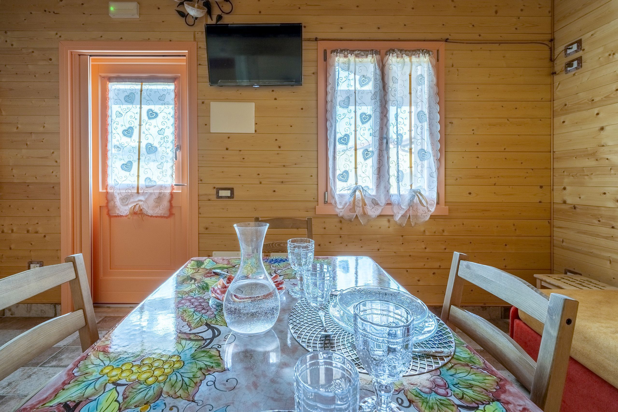 L’intérieur d’un chalet en bois comprend une table à manger pour quatre personnes, un téléviseur fixé au mur et deux fenêtres ornées de rideaux à motifs.