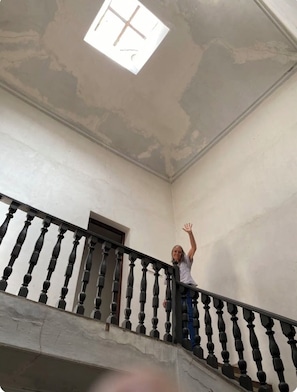 A person standing on a staircase with a black balustrade, a white ceiling with visible plaster marks, and a small window with a cross design.
