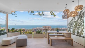 Un balcon moderne, agrémenté d’un canapé, d’une table et de luminaires suspendus, offrant une vue sur le paysage urbain et la mer.