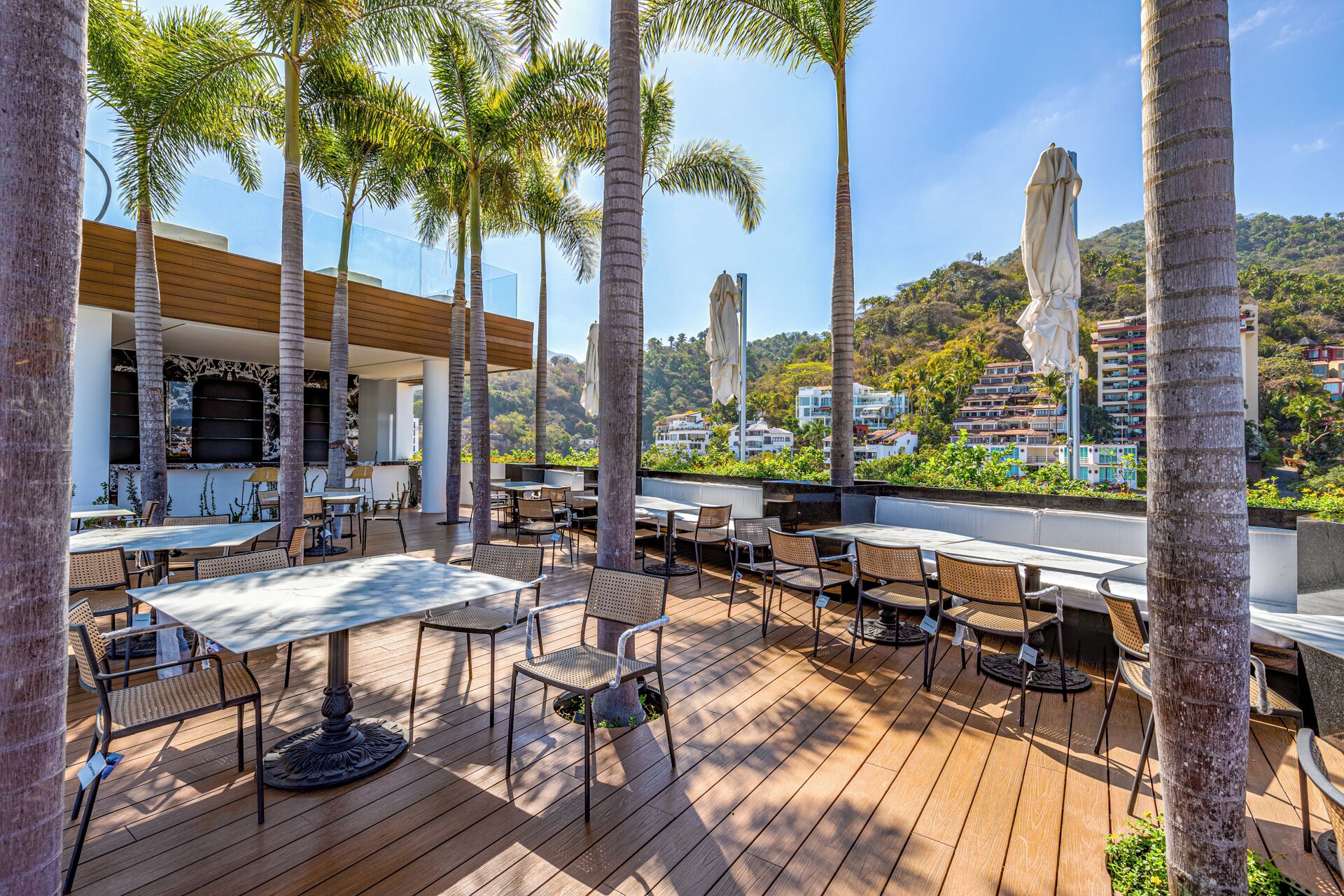Un patio exterior con piso de madera, mesas y sillas, rodeado de palmeras y vistas a una colina con edificios.