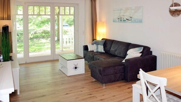 A modern living room with a dark brown sofa, a white table, and a white chair. There is a large window with curtains, a painting on the wall, and a lamp on the table. - Comfy Apartment With 1 Bedroom and Balcony 134 (Binz)