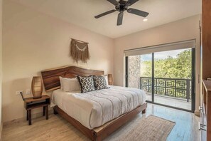 A bedroom with a large bed, a ceiling fan, a nightstand with a lamp, a view through a sliding glass door to an outdoor balcony.