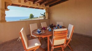 Una mesa redonda de madera con seis sillas, vista al mar a través de una ventana y una planta en maceta sobre el alféizar.