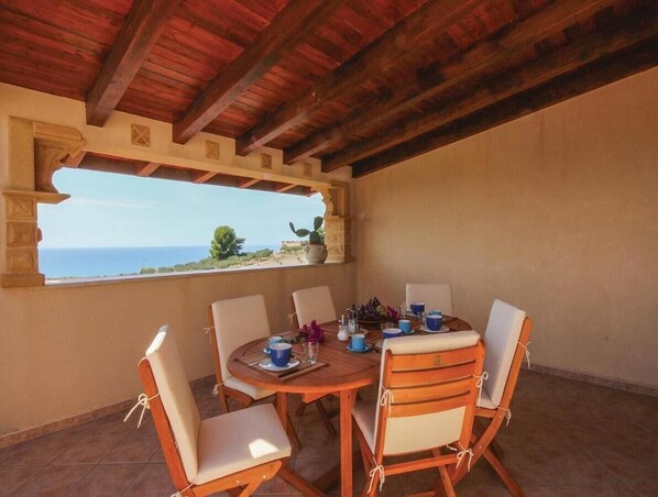 A dining area with a table set for six, surrounded by chairs, under a wooden ceiling with a view of the sea.