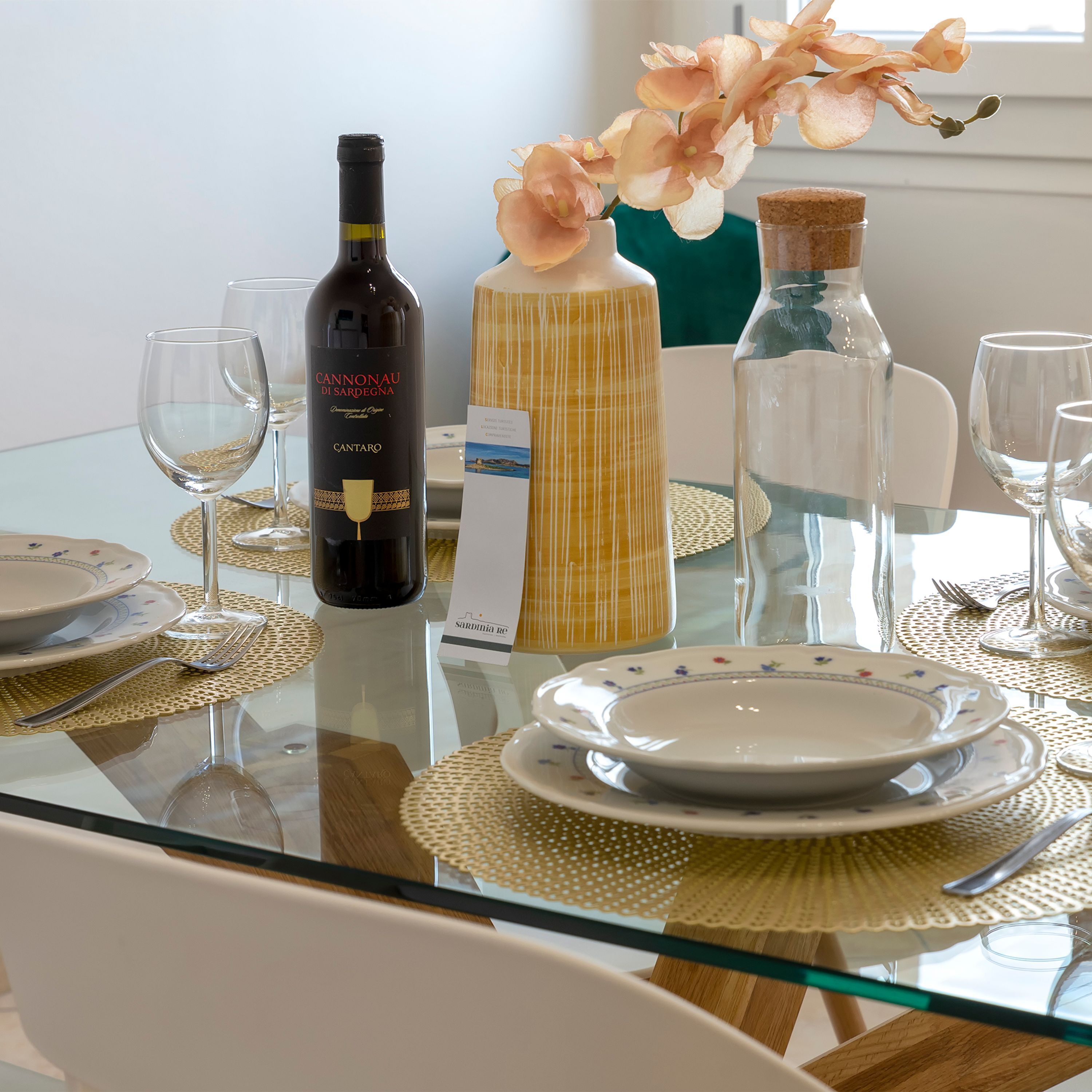 Une table à manger dressée avec des assiettes, des couverts, une bouteille de vin, une bouteille d’eau et un vase rempli de fleurs.