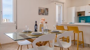A dining area with a glass table, white chairs, and a kitchen in the background. - Sardinia Re - Emerald House (Sassari)
