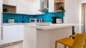 A modern kitchen with white cabinetry, a blue backsplash, and a central island with a sink. There is a yellow chair at the island and a coffee machine on the countertop. - Sardinia Re - Emerald House (Sassari)