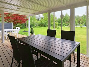 A covered patio with a table and chairs, a small potted plant, and a view of a green lawn and trees. - 6 Person Holiday Home in Idestrup (Idestrup)