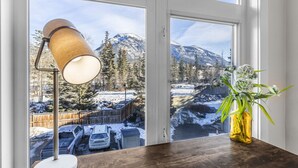 A room with a large window overlooking a snowy landscape with mountains, a wooden table, a lamp, and a vase with flowers.