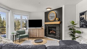 A modern living room with a fireplace, a television, and a view of trees through large windows.