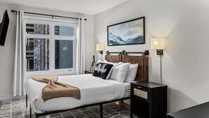 A hotel room with a bed, a nightstand, a lamp, a window with curtains, and a wall-mounted picture.