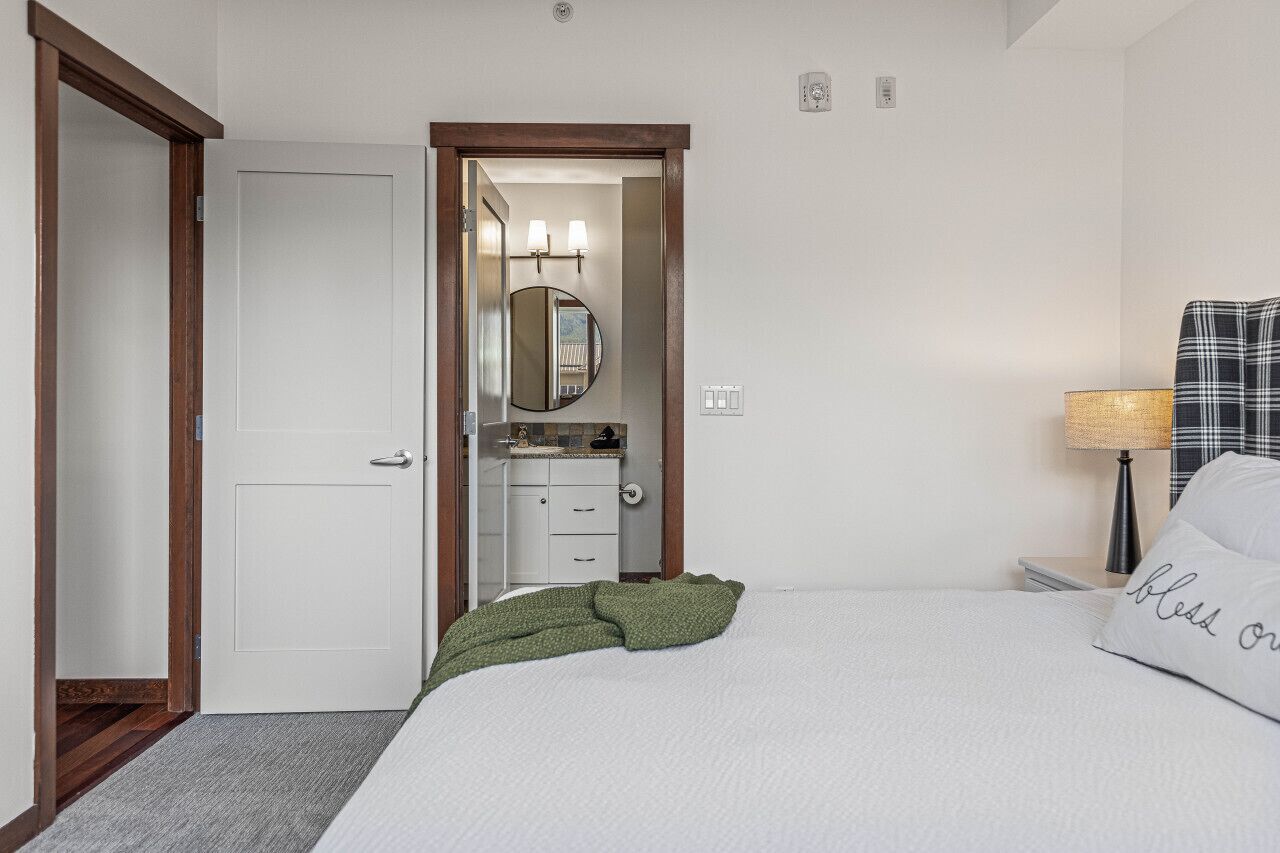 A hotel room with a bed, a bedside lamp, a green throw blanket, a white pillow with text, a bathroom with a mirror and lights, and a door.