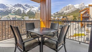 Un balcón con mesa y sillas, con vista a un paisaje montañoso y edificios al fondo.