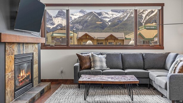 Un salón con chimenea, un ventanal con vistas a la montaña, un sofá gris y un televisor de pantalla plana sobre la chimenea.
