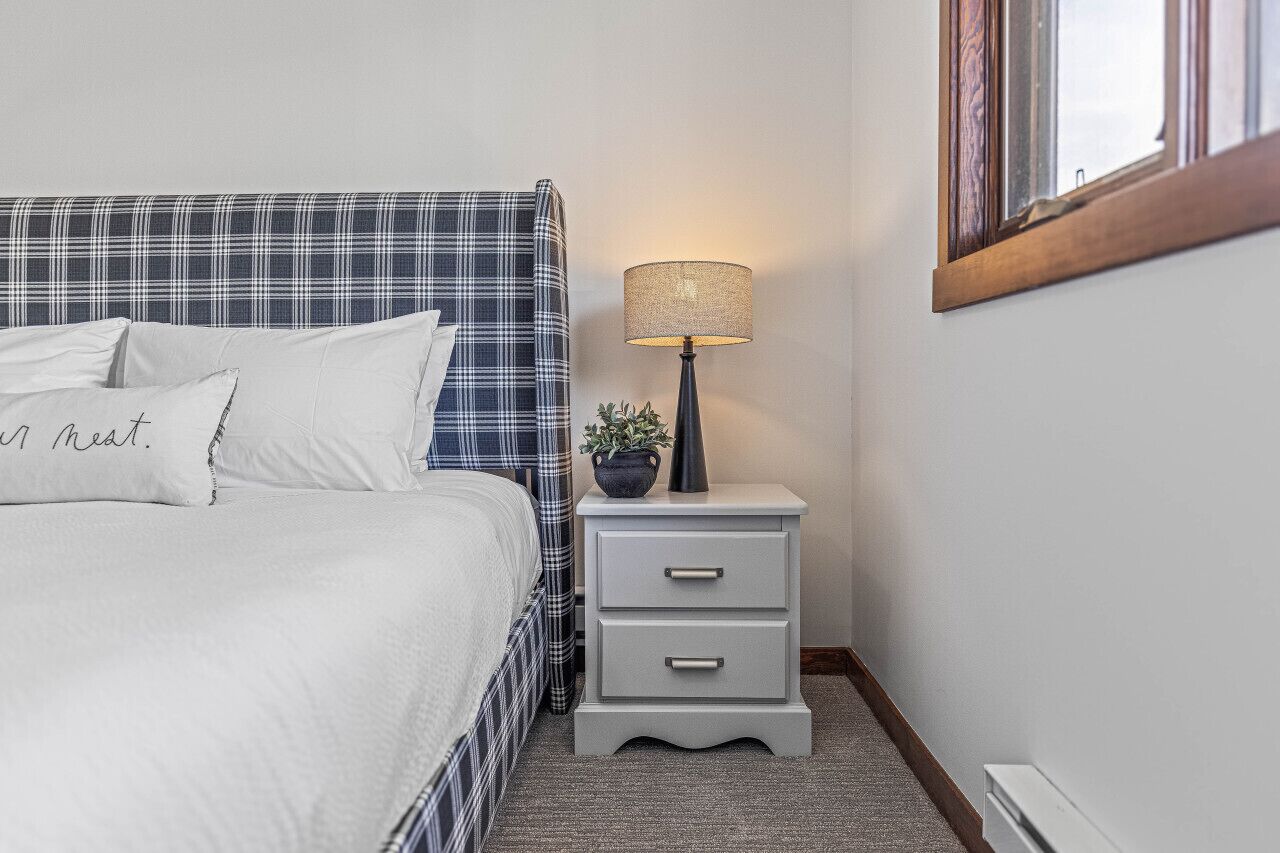 A neatly made bed with a plaid headboard, a bedside lamp, and a small bedside table with drawers.