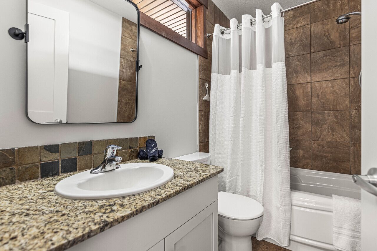 A bathroom with a granite countertop sink, a white toilet, a shower with a white curtain, and a mirror above the sink.