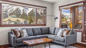 Una sala moderna con un sofá seccional gris, una mesa de centro de madera y un ventanal con vistas a montañas y un edificio.