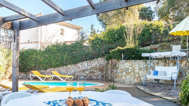 Un espace aménagé au bord de la piscine, avec des chaises longues, une table où sont servis des croissants et du jus d’orange, et un mur en pierre.