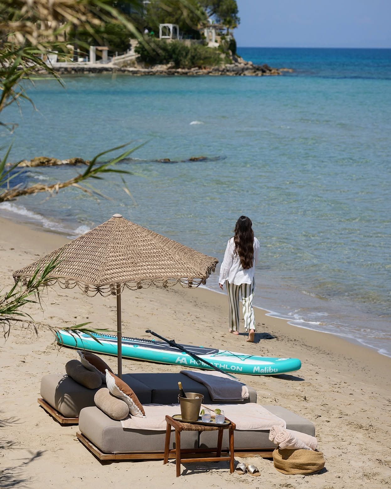Ein Strand mit Liegebereich, einem Stand-Up-Paddleboard und einer Person, die zum Wasser geht.